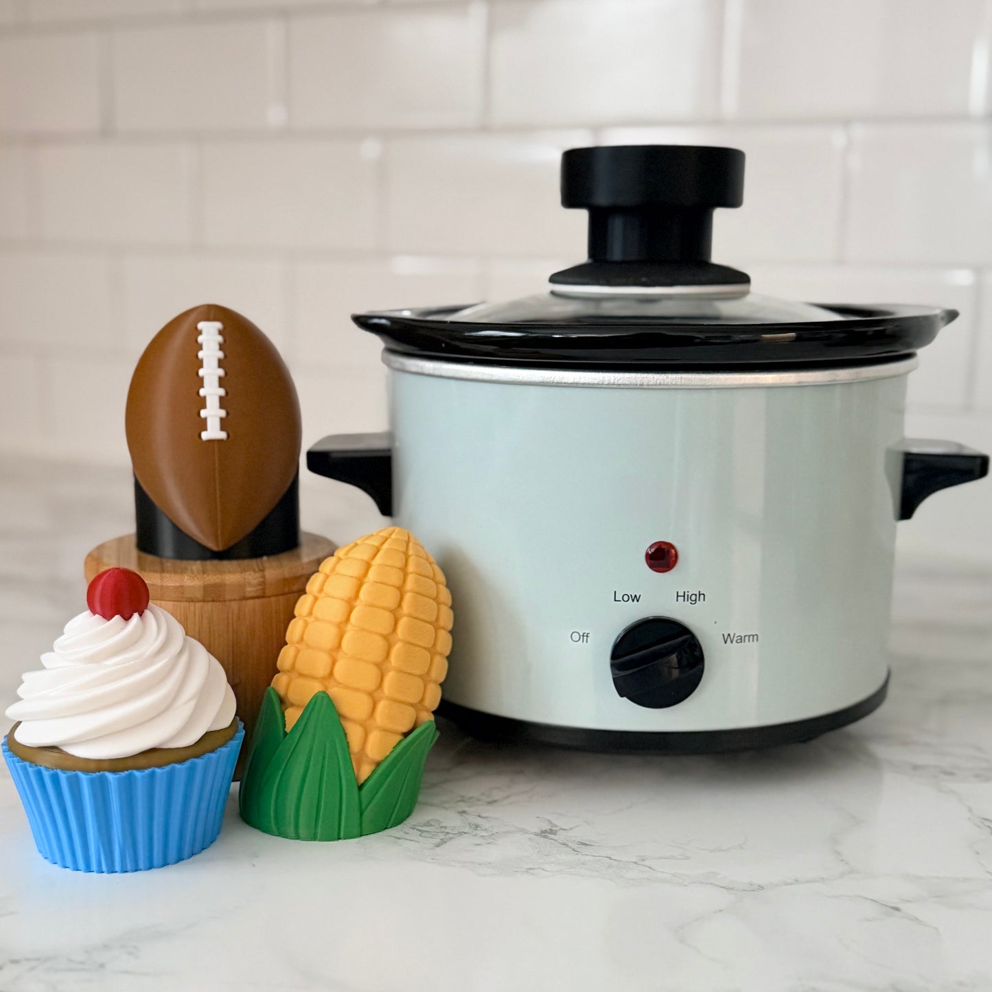 White slow cooker with various crockit handles - american football, cupcake, and corn on the cob.